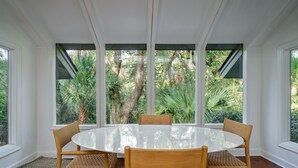 A round glass table with four wooden chairs in a room with large windows overlooking a lush garden.