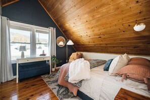 Une chambre confortable avec un plafond en pente, un bureau avec une lampe, un lit avec de nombreux oreillers et une vue par la fenêtre.