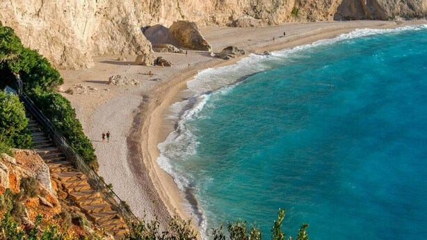 A coastal beach with a rocky cliff, a sandy shoreline, and a few people walking along the beach. - Villa Roya in Sivota (Lefkada)
