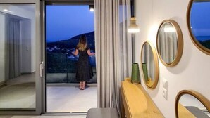 A modern hotel room with a large sliding glass door leading to a balcony, a wooden console table, a gray bench, and a bed. - Villa Roya in Sivota (Lefkada)