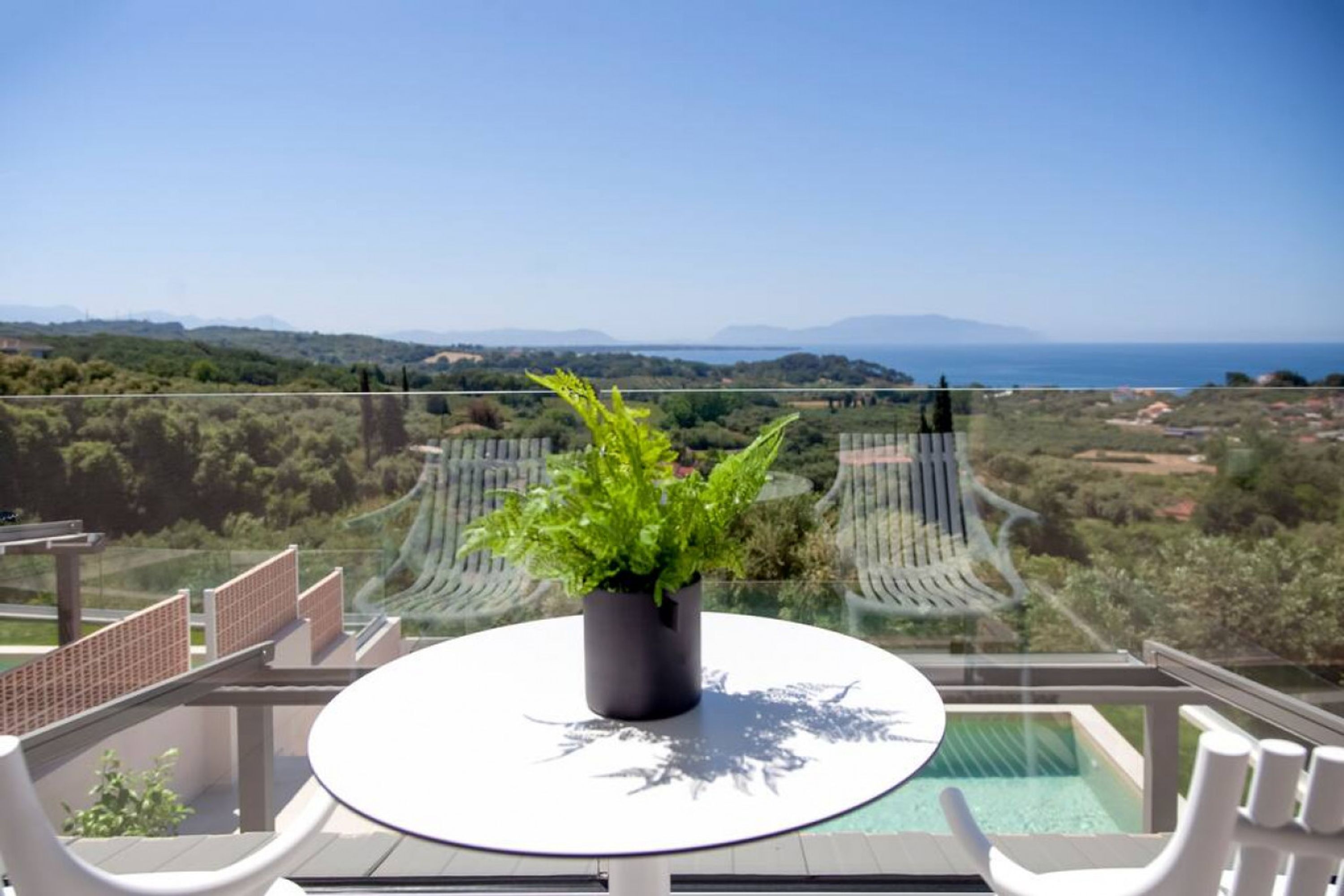 A balcony with a glass table, a potted plant, and a view of the sea and hills.
