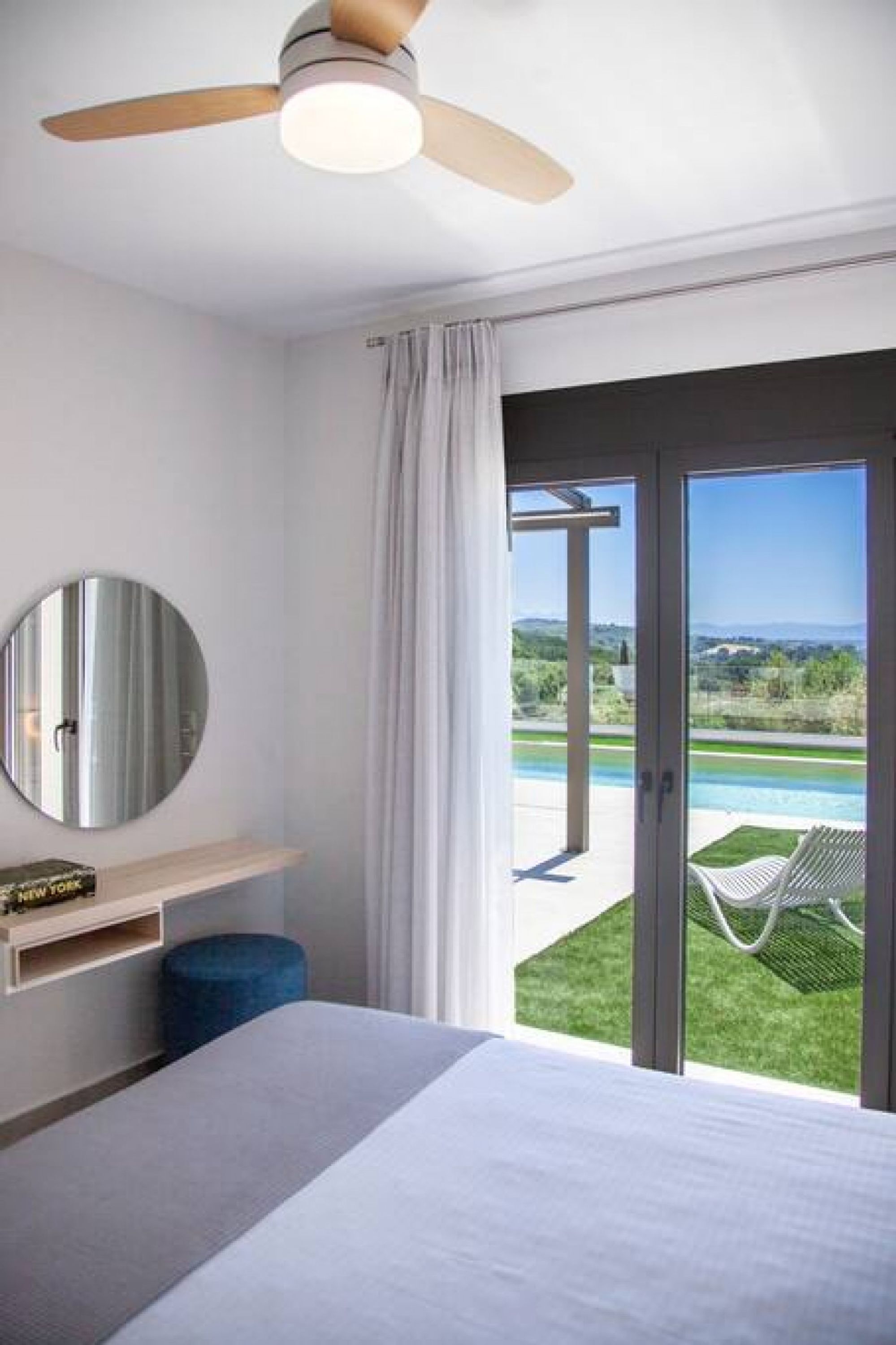 A modern hotel room with a bed, a round mirror, a ceiling fan, a view of a pool and greenery outside.