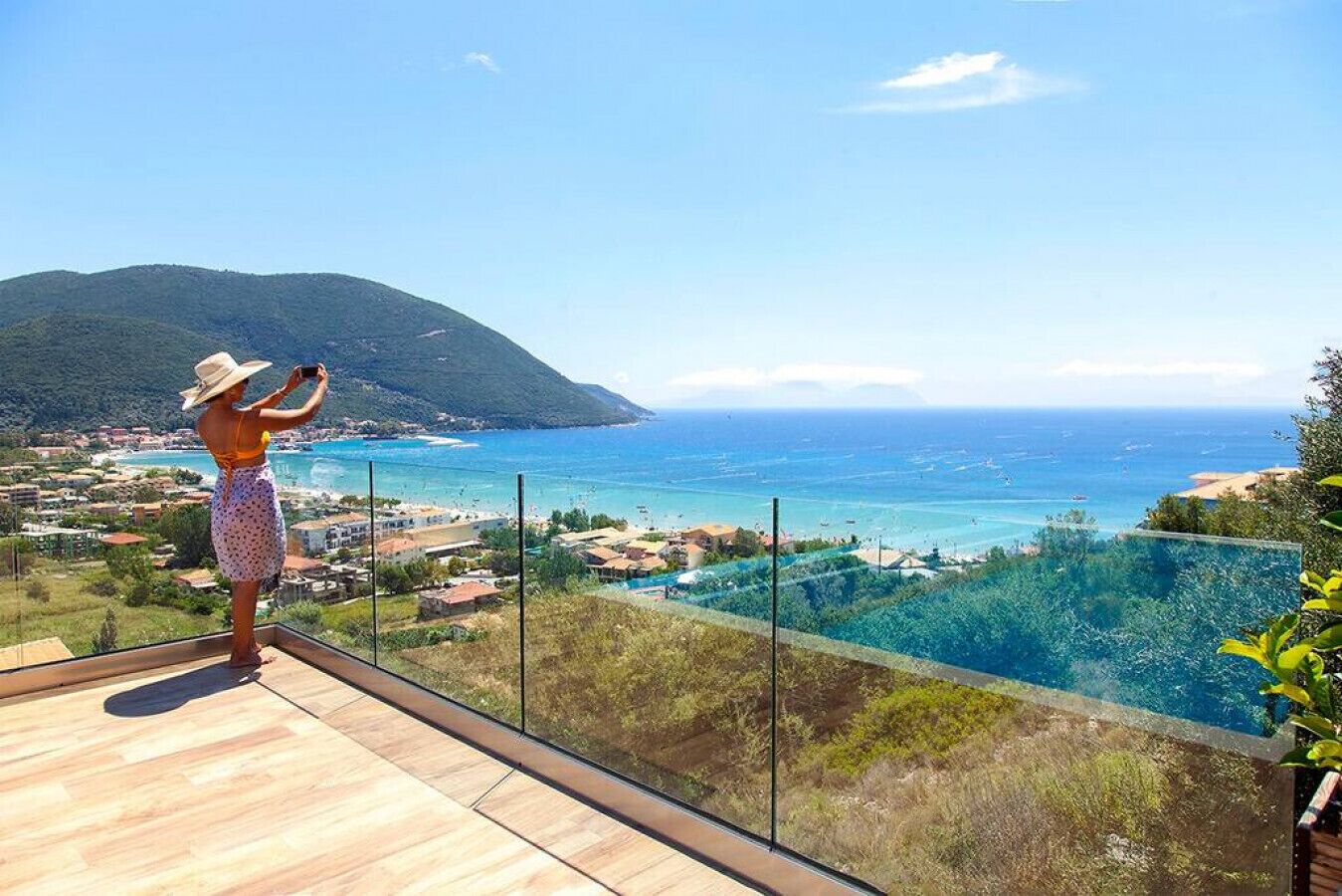 Una persona fotografiando una vista costera desde un balcón con piso de madera y barandilla de vidrio.