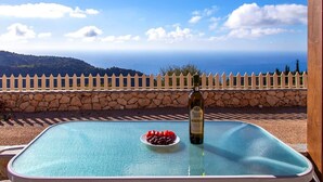 Une table à plateau de verre, sur laquelle sont posés une bouteille de vin et une assiette de raisins, donnant sur un mur de pierres et une clôture blanche en bois, avec une vue sur la mer et les collines.