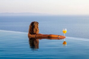 Eine Person entspannt sich an einem Infinity-Pool mit einem Glas Orangensaft und blickt auf ein Gewässer.