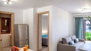 Eine moderne Küchenzeile mit Kühlschrank, Obstschale und zwei Gläsern Orangensaft. Im Wohnbereich sind eine Couch und ein Esstisch zu sehen, mit Blick auf einen Garten durch ein Fenster.