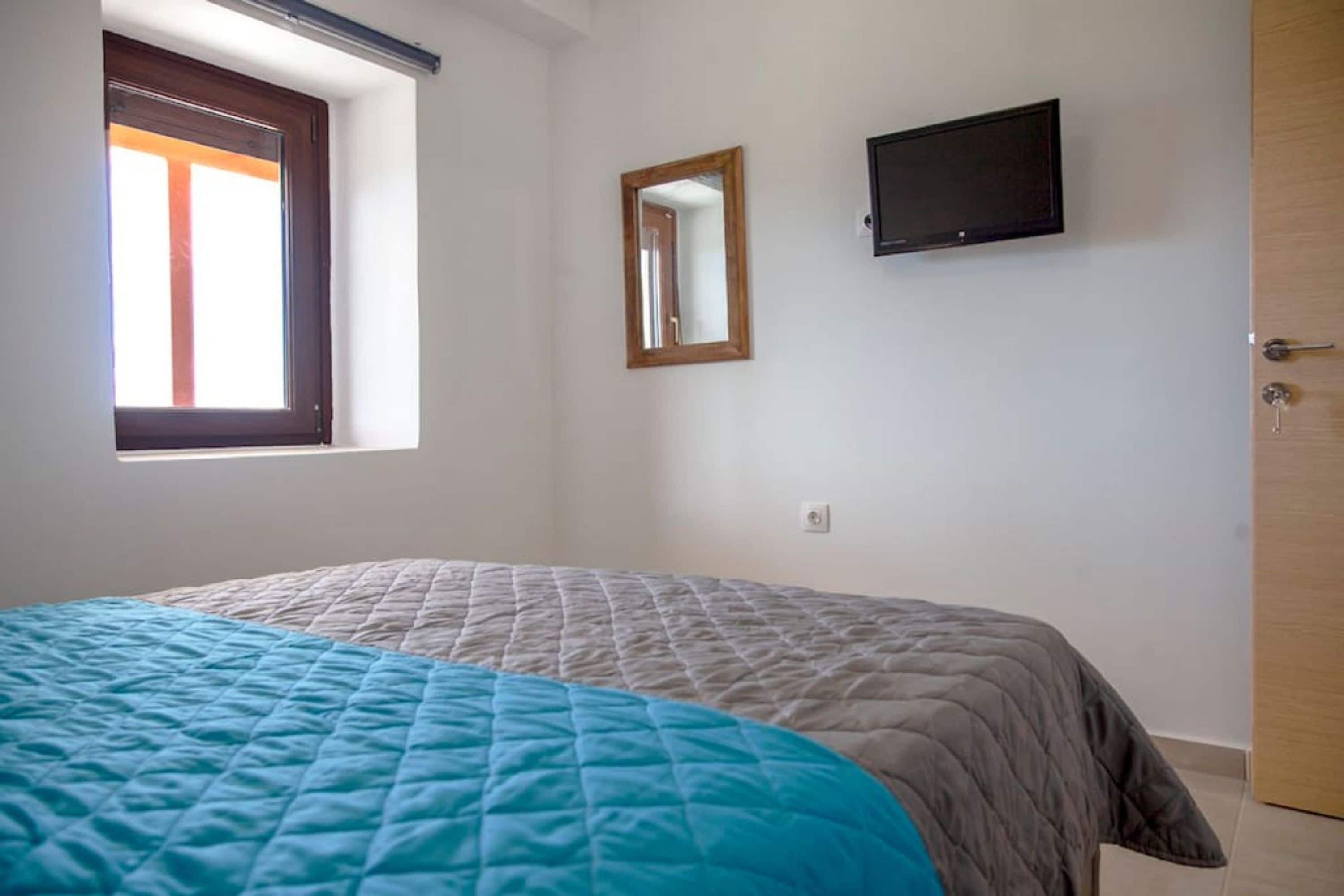 Een slaapkamer met een bed, een raam, een spiegel en een televisie aan de muur.
