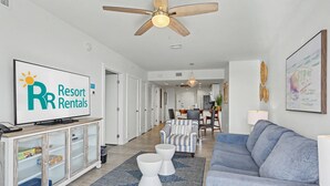 Un salon moderne avec un téléviseur diffusant une émission sur les « locations de vacances », un ventilateur de plafond, un canapé, une table basse et, au fond, un coin repas.