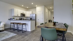 Een moderne keuken met witte kastjes, een magnetron en een koelkast. Er is een eethoek met een houten tafel en groene stoelen.