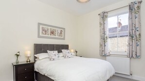 Un dormitorio con cama, mesitas de noche, un cuadro enmarcado y una ventana con cortinas.