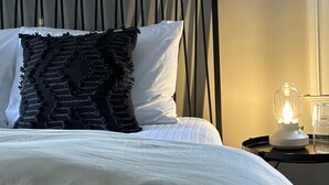 A neatly made bed with white bedding and a black decorative pillow, a bedside table with a glass lamp, and a framed picture of a landscape on the wall.