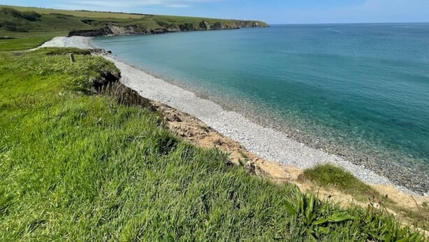 Un paisaje costero con una playa de guijarros, zonas verdes y un cielo azul despejado.