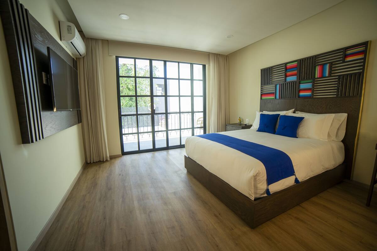 Una habitación de hotel moderna con una cama grande, un televisor de pantalla plana montado en la pared y un ventanal corredero que da a un espacio exterior.