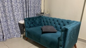 Un canapé recouvert d’un tissu de couleur turquoise, doté d’un dossier capitonné et d’un seul coussin foncé, placé dans une pièce dont le sol est carrelé et où l’on aperçoit, au fond, un rideau à motifs.