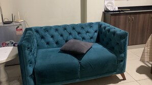 Un canapé recouvert d’un tissu turquoise, agrémenté d’un coussin foncé, dans une pièce où l’on aperçoit un coin cuisine à l’arrière-plan.