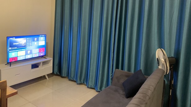 Un salon avec un téléviseur à écran plat posé sur un meuble, un canapé, un ventilateur et des rideaux bleus.