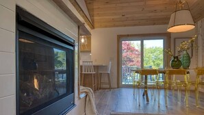 A cozy room with a fireplace, wooden flooring, a dining area with yellow chairs, and a large window with a view of trees.