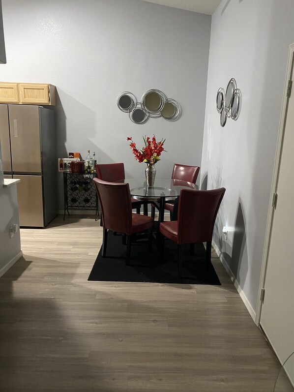 A compact dining area with a round table, four chairs, and a vase of red flowers. A refrigerator and a wine rack are visible in the background.