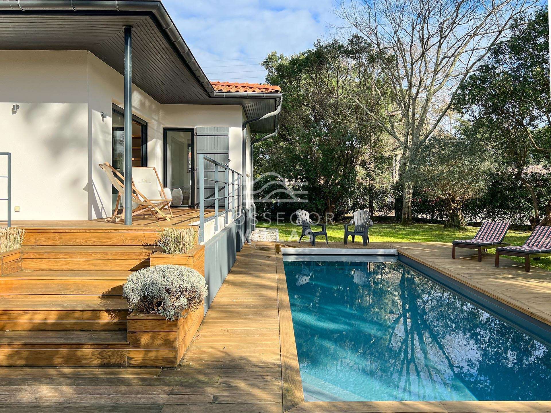 Et moderne hus med swimmingpool, træterrasse og udendørs møbler.