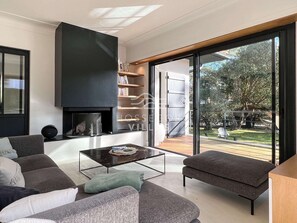 A modern living room with a fireplace, a sofa set, a coffee table, and a large sliding glass door leading to an outdoor area. - Villa XENIA (HOSSEGOR)