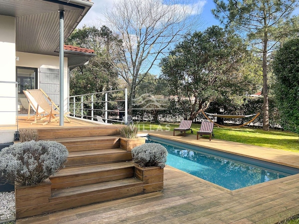 A wooden deck with a pool, outdoor seating, and a view of the surrounding trees.