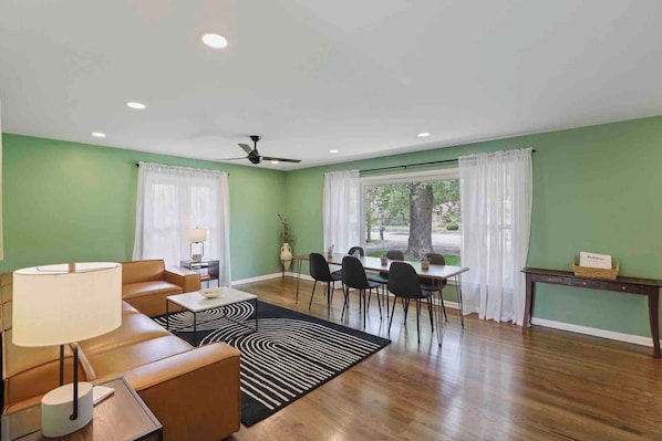 A living room with a sofa, a coffee table, a dining table with chairs, a ceiling fan, and a view of the outdoors through large windows.