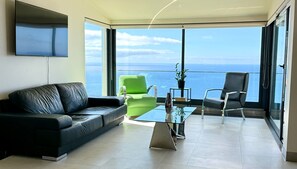 A modern living room with a black leather sofa, a green armchair, and a black chair, all facing a large glass table.