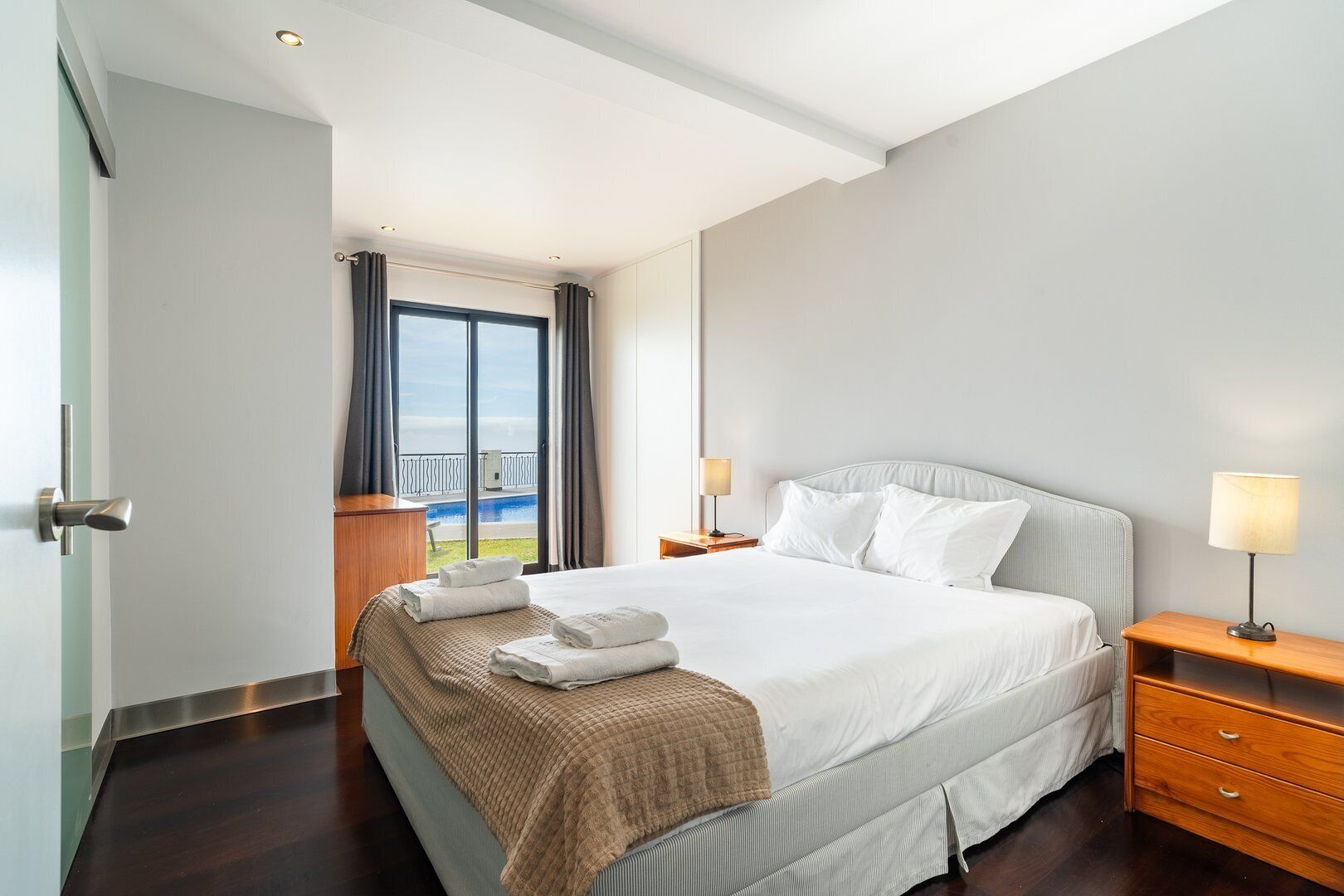 A hotel room with a large bed, a wooden nightstand, a lamp, and a view of the sea through a large window.