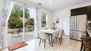 A modern kitchen with a stainless steel refrigerator, a dining table with chairs, and a sliding glass door leading to a patio with a chair and a flower vase.