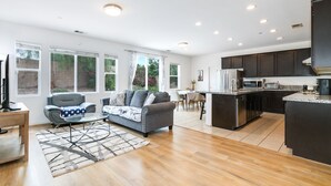 A modern living room with a grey sofa, a coffee table, and a TV on a wooden stand. There is a kitchen area with dark cabinets and a microwave.