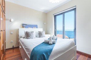 Une chambre à coucher avec un lit, une serviette bleue, une tête de lit blanche, une fenêtre donnant sur la mer et une armoire en bois.