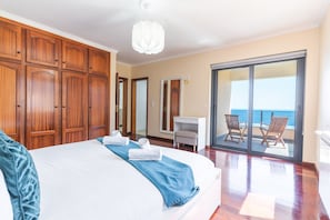 A bedroom with a large bed, a desk, a chair, and a balcony with a view of the sea.