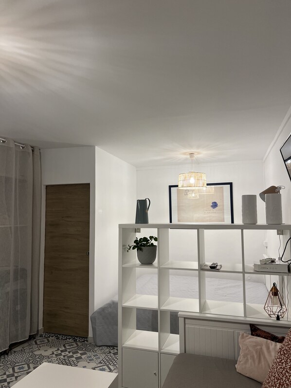A modern bedroom with a white bed, a white shelving unit, a potted plant, a framed picture, and a hanging light fixture.