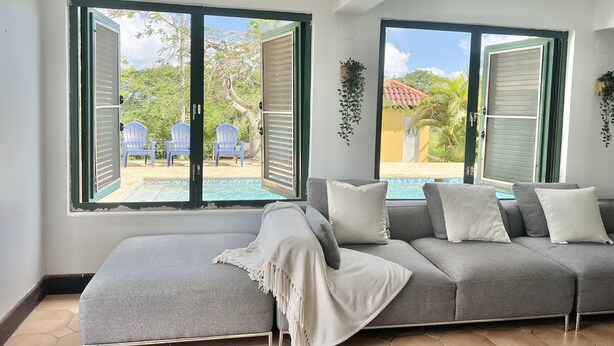 Una sala de estar moderna con un sofá gris, almohadones blancos y vistas a una piscina y sillas de exterior a través de amplias ventanas.