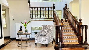 Un amplio salón con escalera, una araña, un sillón estampado y una pequeña mesa con libros y una planta en maceta.
