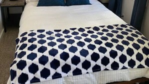 A neatly made bed with a patterned bedspread, two bedside lamps, a painting above the bed, and a window with curtains. - Hyde Park Hyde Out Apt 3 (Boise)
