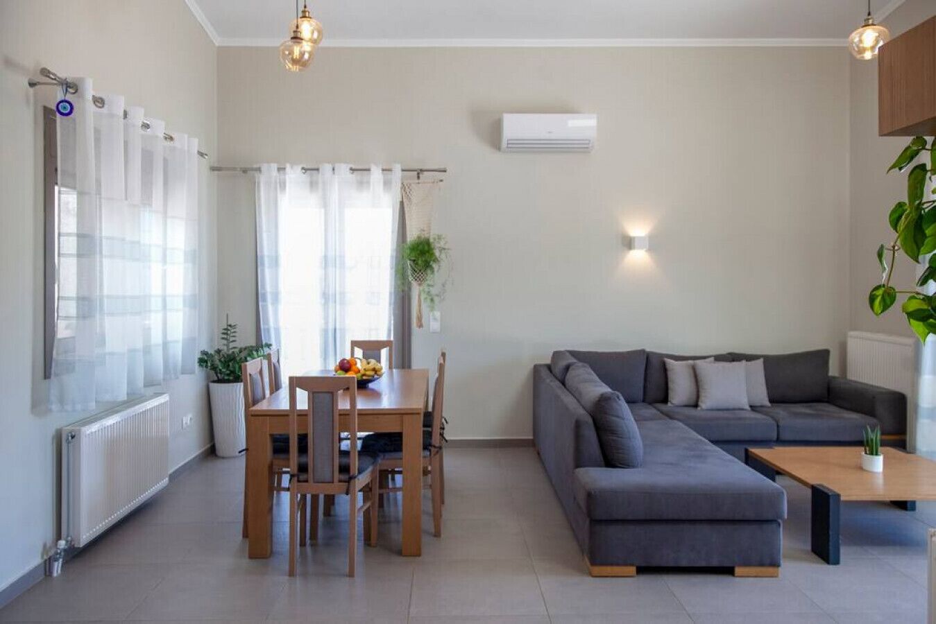 Una sala moderna con un sofá gris, una mesa de comedor de madera y sillas. Se ve un radiador, una ventana con cortinas traslúcidas y un aire acondicionado de pared.