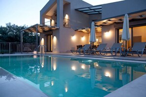 A modern pool area with lounge chairs and umbrellas, surrounded by a building with large windows and a balcony. - Villa Alkistis in Lefkada (Lefkada)