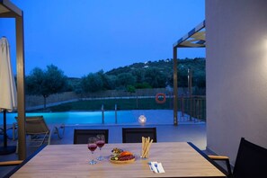 A dining area with a table set for two, a pool in the background, and a view of a hillside. - Villa Alkistis in Lefkada (Lefkada)