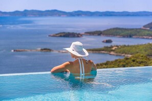 Una persona relajándose en una piscina infinita de color azul cristalino, con vistas panorámicas del mar y de islas lejanas.