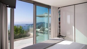 Une chambre moderne dotée d’une grande porte-fenêtre coulissante donnant sur un balcon avec vue sur la mer et les montagnes.