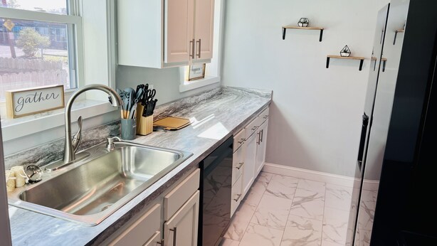 A modern kitchen with a marble countertop, stainless steel sink, and white cabinetry. There are two wooden cabinets above the countertop and a black refrigerator on the right. - Remodeled townhouse - 5 mins from Shawnee Sports Complex! (Dunbar)