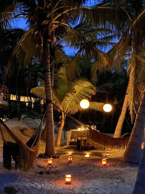 A hammock under palm trees with a table and chairs, surrounded by sand and lit candles. - CASA NALUM (Tulum)