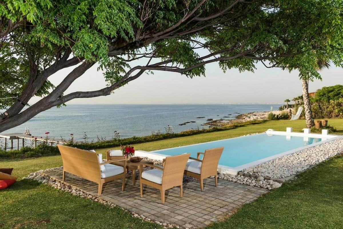 A pool area with a view of the sea, surrounded by greenery and a patio with chairs.