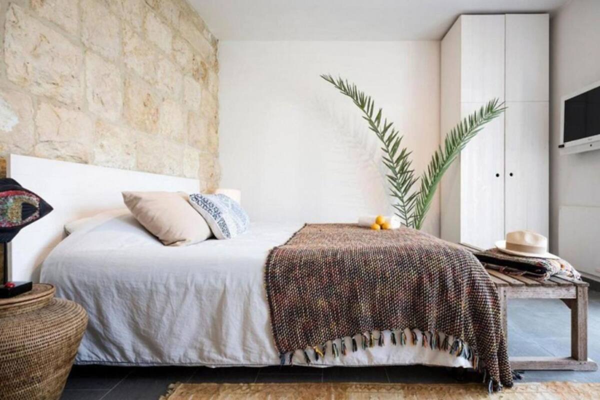 A bedroom with a stone wall, a bed with a white headboard, a woven basket, and a wooden bench with a hat.