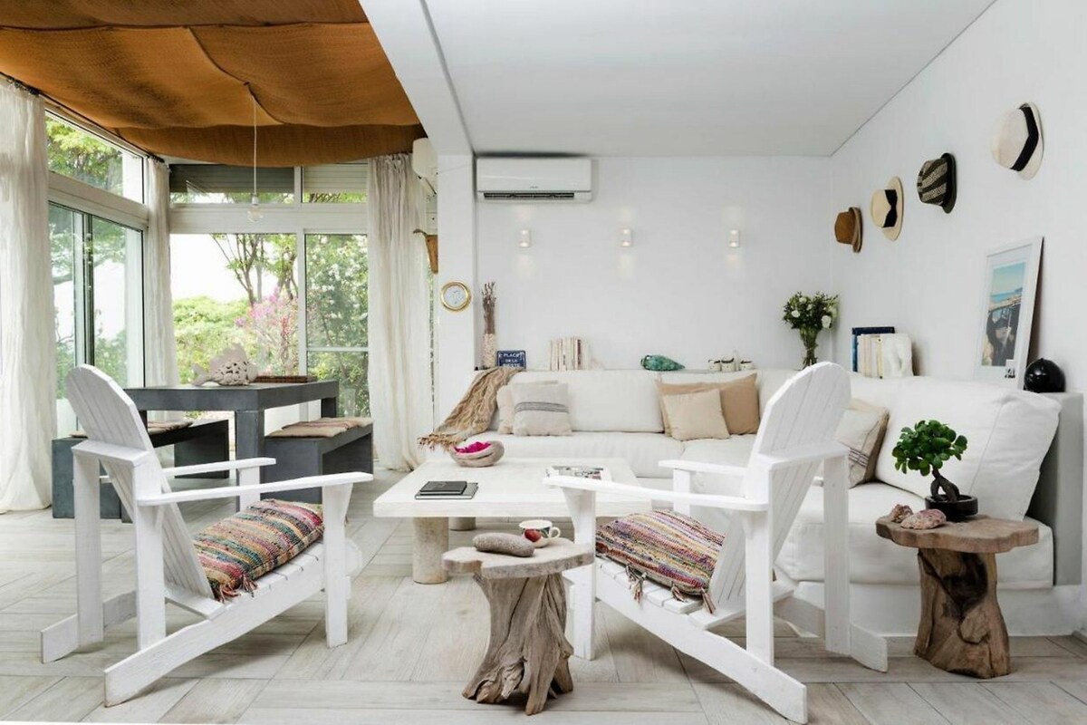 A modern living room with a white sofa, two white chairs, a coffee table, and a dining table. There are decorative items on the walls and a window with a view of greenery.