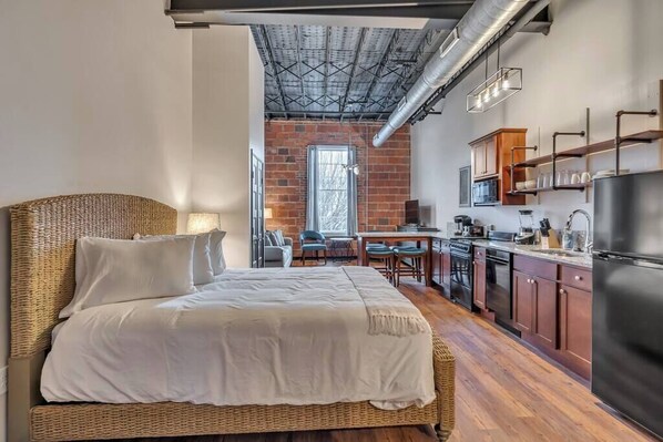 A room with a bed, kitchenette, and a brick wall.