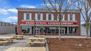 A brick building with a sign reading 'Grand Strand Brewing Company.' There are outdoor seating areas with chairs and tables. - 1 Bedroom @ Grand Strand Brewery 203 (Myrtle Beach)