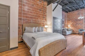 A bedroom with a brick wall, a bed with a wicker headboard, a grey sofa, and a desk with a chair. - Beach Brews & Coastal Views Loft (Myrtle Beach)