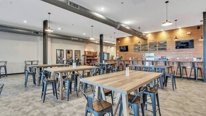 A spacious indoor area with multiple wooden tables and blue chairs, a kitchen area with a counter, and a wall-mounted TV.